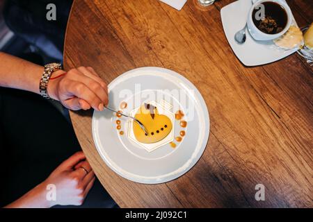 Serviert Tisch mit Speisen und Getränken, Teller mit herzförmigen Kuchen mit Glasur neben einer Tasse Kaffee, Essen im Restaurant, Person Hand dekoriert Stockfoto