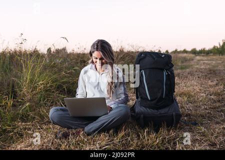 Junge Frau mit einem Rucksack neben ihr, die mitten auf dem Feld einen Laptop für die Telearbeit benutzt. Digitaler Nomade. Stockfoto