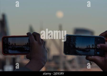 London, Großbritannien. 17. März 2022. Wetter in Großbritannien – die Menschen fotografieren den Vollmond, der über der City of London aufgeht. Der Vollmond dieses Monats wird laut Almanach des Alten Bauern als Wurmmond bezeichnet, wenn Regenwürmer erscheinen, während sich der Boden im Frühjahr erwärmt. Es wird auch so von indianischen Stämmen benannt, die sich auf eine andere Art von „Wurm“ beziehen - Käferlarven - die zu dieser Zeit aus der tauenden Rinde von Bäumen und anderen Winterverstecken auftauchen. Kredit: Stephen Chung / Alamy Live Nachrichten Stockfoto