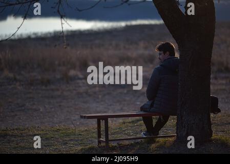 Mann, der auf einer Bank bei einem Baum sitzt. Frühfrühlingshafte Natur und Sonnenuntergang Stockfoto