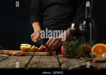 Nahaufnahme des Küchenchefs, der Apfel auf Holzbrett schneidet, bevor er Glühwein auf einem rustikalen Holztisch mit festlichem Hintergrund zubereitet. Backstage von c Stockfoto