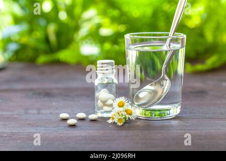 Homöopathisches Konzept. Trinkglas mit Wasser, Dragees und Kamille auf einem Holztisch. Leuchtendes Grün im Hintergrund. Fokus auf den Vordergrund. Stockfoto