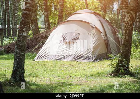 Ein großes Familienzelt wird auf weichem grünen Gras im Wald in der Nähe des Sees aufgestellt. Aktivitäten am Wochenende und Camping-Abenteuer Stockfoto