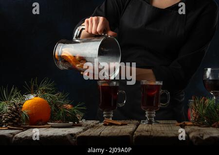 Der professionelle Koch gießt Glühwein in Gläsern auf einen rustikalen Holztisch mit festlicher Komposition. Backstage beim Kochen eines heißen Getränks mit Duft Stockfoto