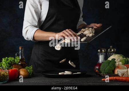 Der professionelle Koch in schwarzer Schürze gießt gehackte Pilze auf den Teller, um Suppe auf dunkelblauem Hintergrund zu kochen. Backstage der Zubereitung der Mahlzeit. Gesund Stockfoto
