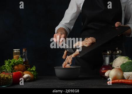 Der professionelle Koch in schwarzer Schürze gießt Hühnerfilet in die Bratpfanne, um Suppe auf dunkelblauem Hintergrund zu kochen. Backstage der Zubereitung der Mahlzeit. Gesund Stockfoto