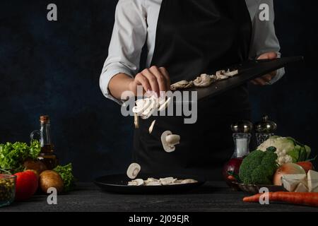 Nahaufnahme des Küchenchefs in schwarzer Schürze, der gehackte Pilze auf den Teller gießt, um Suppe auf dunkelblauem Hintergrund zuzubereiten. Backstage der Zubereitung der Mahlzeit. Gesund Stockfoto