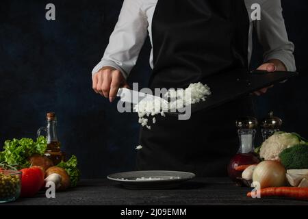 Der professionelle Koch in schwarzer Schürze gießt gehackte Zwiebeln auf den Teller, um Suppe auf dunkelblauem Hintergrund zuzubereiten. Backstage der Zubereitung der Mahlzeit. Gesund d Stockfoto