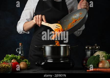 Der Profi-Koch gießt Frittiergemüse in die Pfanne, um Suppe auf dunkelblauem Hintergrund zuzubereiten. Backstage der Zubereitung der Mahlzeit. Gesundes Gericht. Lebensmittelkonzept. Stockfoto