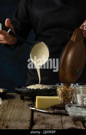 Makroaufnahme des Prozesses der Teiggiessen auf Waffelmaschine für die Zubereitung süßer Waffel auf rustikalem Holztisch mit Zutaten auf dunkelblau. Fro Stockfoto