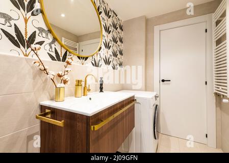 Geflieste Toilette mit großen cremefarbenen Fliesen, goldenem Wasserhahn, ägyptischer Baumwollzweig und dekorativem Papier, weißem Aluminium-Handtuchheizkörper und weißer Waschmaschine Stockfoto