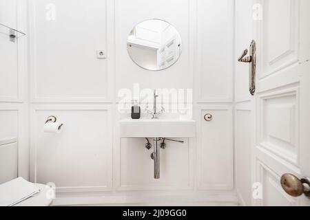 Hängendes Waschbecken aus weißem Porzellan an einer weißen Holzwand mit Vintage-Wasserhahn und rundem Spiegel Stockfoto