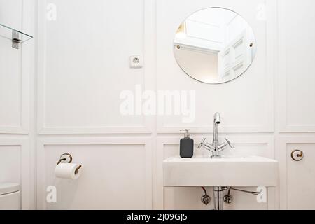 Weißes Porzellanwaschbecken an einer weißen Wand mit dekorativen Chrom-Akzenten, einem runden rahmenlosen Spiegel, einer Seifenschale aus schwarzem Glas Stockfoto