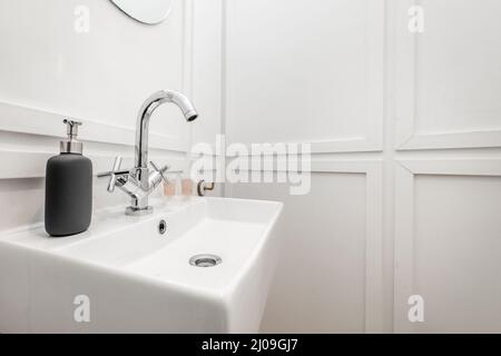 Kleines weißes Porzellanwaschbecken, das an einer weißen Wand mit dekorativen Chromakzenten in einer Ferienwohnung hängt Stockfoto