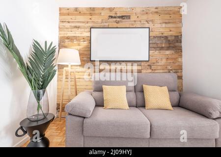 Wohnzimmer mit einer mit Brettern bedeckten Wand, einem Palmenblatt in einer Glasvase, einem grauen Zweisitzer-Sofa und einem Teppich aus Naturfaser, einer Stativlampe und weißem Schmerz Stockfoto