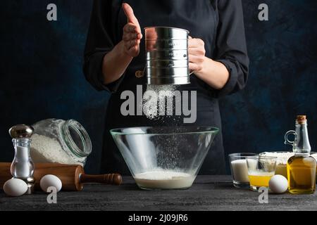 Der Profi-Koch in schwarzer Uniform siebt Mehl in die Glasschüssel, um den Teig zuzubereiten. Zutaten für das Kochen auf dem Hintergrund. Dunkelblau b Stockfoto