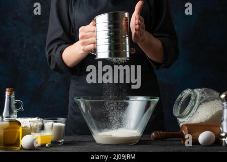 Makroaufnahme des Küchenchefs in schwarzer Uniform siebt Mehl in die Glasschüssel für die Zubereitung des Teiges. Zutaten für das Kochen auf dem Hintergrund. Dunkelblauer Rücken Stockfoto