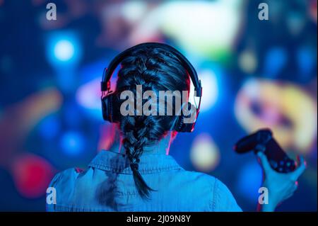 Blick von der Rückseite der Gamer Frau mit Pigtail spielen Videospiel zu Hause vor einem großen Bildschirm mit Kopfhörer. Bunte Neon LED Lichter Hintergrund. Stre Stockfoto