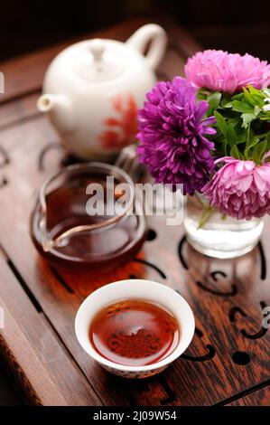 Kleines Bouquet aus lila und rosa Astern und schwarzem Tee in chinesischer Tradition auf geschnitztem Tee Tablett vertikal Stockfoto