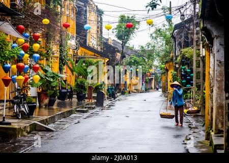 Eine Vietnamesin, die mit ihrem Polenträger eine nasse Straße in der Altstadt entlang geht. Stockfoto