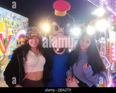 Zwei Latina-Frauen, die bei einem Nachtmarkt neben einem Schauspieler stehen, der sich wie ein Hund kleidet Stockfoto