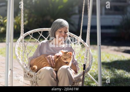 Ältere Frau, die im Hinterhof eine Ingwerkatze auf dem Rollstuhl hält. Stockfoto