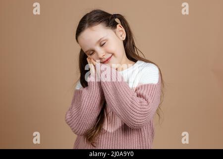 Portrait eines jungen ehrfürchtig lächelnden Mädchens mit langen dunklen Haaren in weißem, rosa Pullover, stehend mit geschlossenen Augen, liegend auf gefalteten Händen unter der Wange Stockfoto