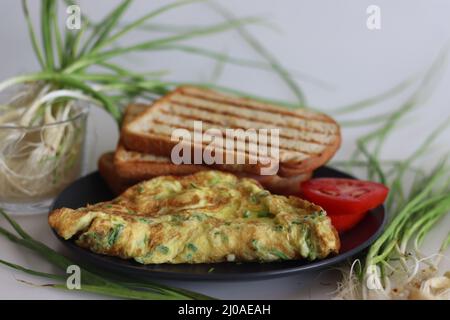 Eieromelett mit fein gehacktem grünem Knoblauch, grünen Chilis mit Butter, geröstetem Brot und frischen Tomaten serviert. Aufgenommen auf weißem Hintergrund. Stockfoto