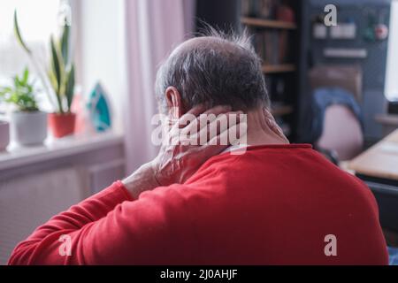 Ich brauche eine Massage. Frustrierter älterer Mann in rotem Pullover, der die Hand auf dem Nacken hält und auf dem Sofa sitzt Stockfoto