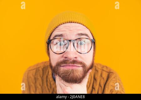 Schöner junger bärtiger Kaukasischer Mann in Hut und Brille zeigt Interesse Blick auf die Kamera mit großen Augen berühren seinen Bart Hipster Headshot isoliert selektiven Fokus in den Kopf orange Hintergrund Studio-Aufnahme . Hochwertige Fotos Stockfoto