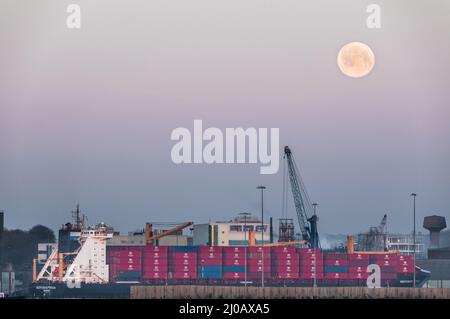 Ringaskiddy, Cork, Irland. 18.. März 2022. Ein Wurm Moon desends als Containerschiff Independent Vision lädt mit einer Ladung nach Chester, USA, in Ringaskiddy Docks, Co. Cork, Irland. - Credit; David Creedon / Alamy Live News Stockfoto