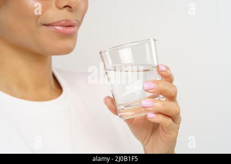 Junge afrikanerin hält Glas mit reinem Mineralwasser, während sie isoliert über der grauen Wand steht Stockfoto