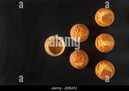 Stillleben mit einer Draufsicht auf eine Gruppe von Cupcakes mit einem der leeren Behälter, in einer dreieckigen Form, auf einem dunklen Hintergrund platziert. Stockfoto