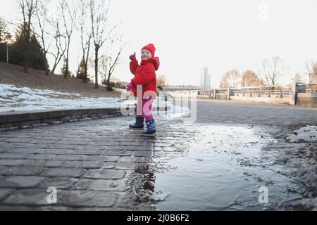 Happy Baby Mädchen in Stiefeln und rote Jacke laufen und spielen in Wasserpfütze. Lustige fröhliche Kleinkind Mädchen im Freien spielen Stockfoto