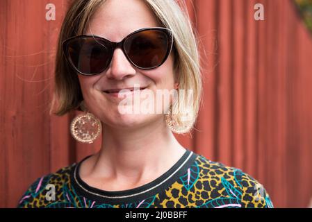 Porträt einer attraktiven dreijährigen Frau mit Sonnenbrille, die an einer typischen roten Falun-Wand steht, Falun, Dalarna, Schweden Stockfoto