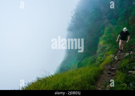 Weißer Mann mit schwarzem Hemd und Hut, der über den Wolken auf dem Berg auf Kap verde entlang einer tiefen nebligen Klippe geht Stockfoto