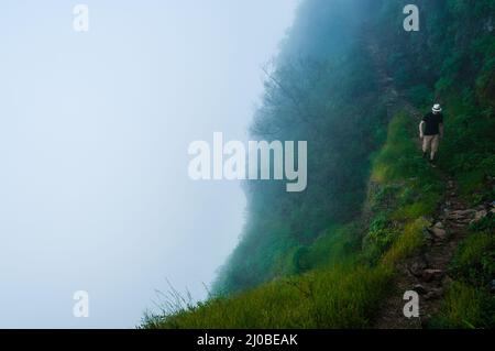 Weißer Mann mit schwarzem Hemd und Hut, der über den Wolken auf dem Berg auf Kap verde entlang einer tiefen nebligen Klippe geht Stockfoto