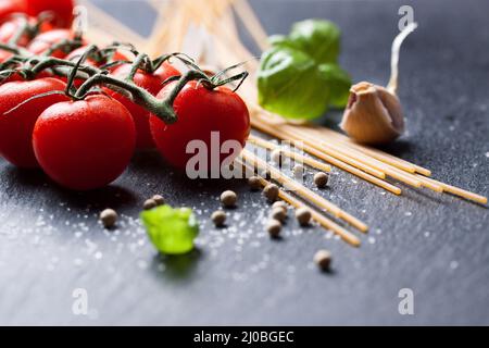 Pasta-Zutaten. Kirschtomaten, Spaghetti, frisches Basilikum, Gewürze auf dunklem Steingrund, Stockfoto
