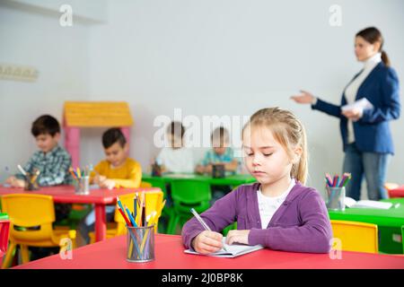 Nettes präteen Mädchen studieren im Klassenzimmer auf Hintergrund mit Klassenkameraden und Lehrer Stockfoto