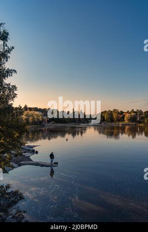 Ein Fischer in der Abenddämmerung fischt, steht auf einer Felsspalte, die das Wasser durchdringt. Im Hintergrund ist ein Streifen von Bäumen, die vom letzten Licht des Tages erleuchtet sind. Hochwertige Fotos Stockfoto