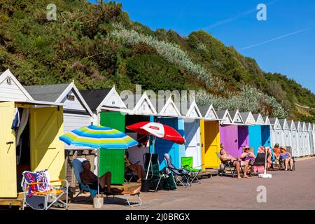 England, Dorset, Bournemouth, Bournemouth Beach, Menschen, die vor einer Reihe von farbenfrohen Strandhütten sonnenbaden Stockfoto