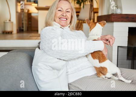 Glückliche Rentnerin, die zu Hause auf dem Sofa mit ihrem Hund auf dem Schoß sitzt Stockfoto