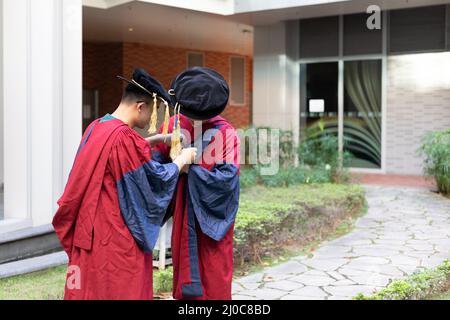 Zwei glückliche, stolze Doktoranden helfen sich gegenseitig, sich akademisches Kleid oder Kleid anzuziehen Stockfoto