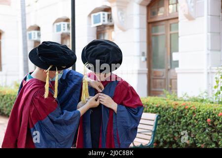 Zwei glückliche, stolze Doktoranden helfen sich gegenseitig, sich akademisches Kleid oder Kleid anzuziehen Stockfoto