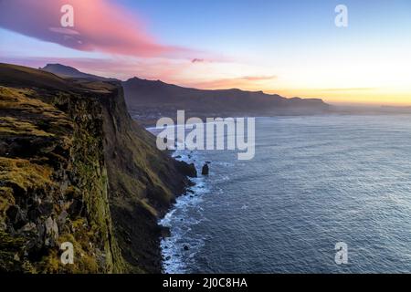 Tagesanbruch entlang der Südküste Islands, mit zerklüfteten, moosbedeckten Klippen und Steinmeersteinen. Die Stadt Vik ist eingebettet in die Bucht zu sehen. Stockfoto