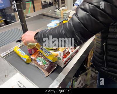 Karlsruhe, Deutschland - 12. März 2022: Seitenansicht des Mannes, der mehrere Lebensmittel arrangiert Süßigkeiten Gemüseobst Konfitüren auf dem Ladentisch im Supermarkt - Lebensmitteleinkäufe Stockfoto