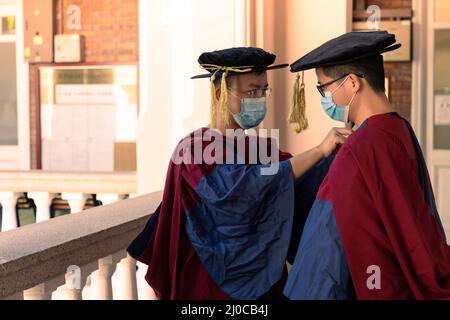 Zwei glückliche, stolze Doktoranden helfen sich gegenseitig, sich akademisches Kleid oder Kleid anzuziehen Stockfoto