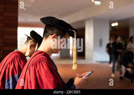 Zwei glückliche stolze Doktoranden im akademischen Kleid lesen Mobiltelefon Stockfoto