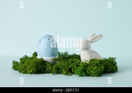 Osterkomposition mit weißem Porzellankaninchen und blauem Ei auf blauem Hintergrund. Grußkarte. Konzept Eiersuche. Stockfoto