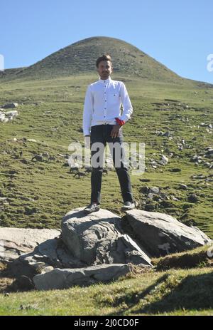 Ein hübscher junger indischer Mann mit weißem Hemd und schwarzem jean, der auf die Kamera schaut, während er über dem Felsen im Berg steht.
